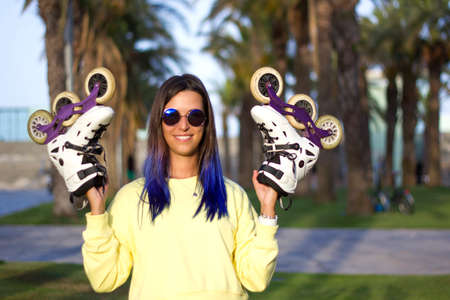 Young smiling informal girl with blue hair and retro sunglasses holding roller skates in her hands....