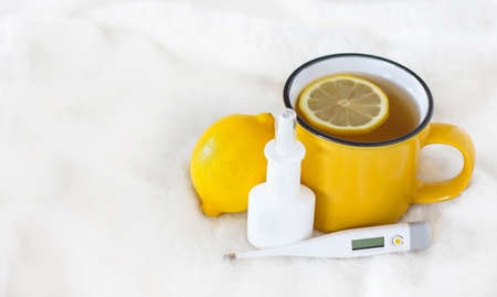yellow cup of tea with lemon next to nasal spray and thermometer on light background with space for...