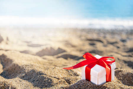 White gift box with a red ribbon standing on the sand with blue sea and sun on background. Holidays on the beach next to the sea.の写真素材