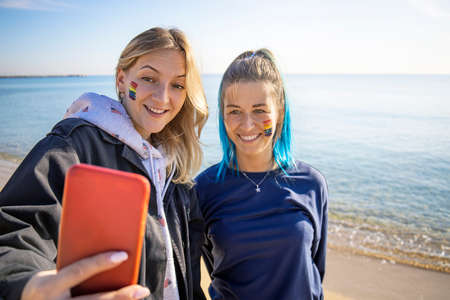 Two happy LGBT girlfriends taking selfie on the beach. Rainbow homosexual flag sign on face