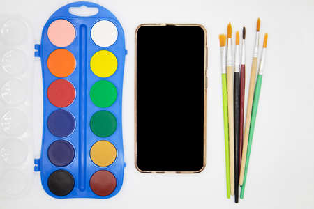 Remote drawing lesson on mobile phone using Internet technologies. E-learning. Top view. Painting online courses. Paints, smart phone with empty blank screen and brushes on white backgroundの写真素材