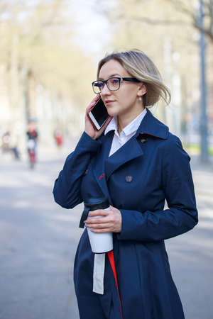 Confident businesswoman using smart phone on the street. Blond young woman with glasses, coffee cup Talking On Phone Walking On Street. Female Business Style. Stylish Business Woman Near Office.の写真素材