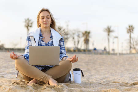 Casual young woman meditating on the empty beach with laptop and coffee cup. Freelance, time management concept. Stress at Work. Space for text.の写真素材