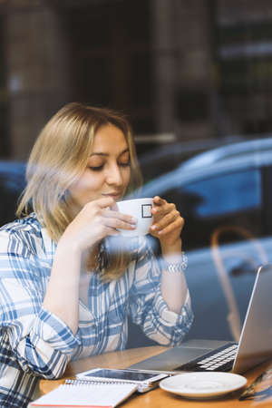 A young blonde woman sitting in cafe with cup of coffee and laptop. View through the window. Telework and freelance. Woman working or online course in a cafe. Woman sniffing and enjoying coffeeの写真素材