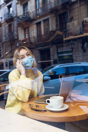Young blond woman with a face medical mask talking mobile phone and working in cafe. Freelance and...