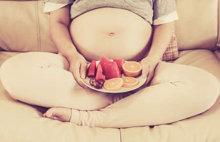 Pregnant woman sitting on couch and holding a plate wirh fruitの写真素材