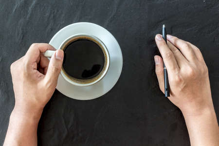 Hand holding cup of coffee and pen on black backgroundの写真素材