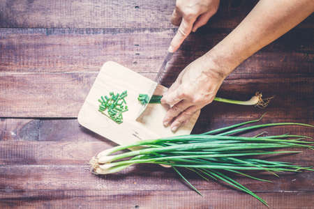 Hand slicing green onions with Knife on chopping board on wooden background