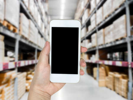 Hand holding mobile smart phone on shelf in Warehouse blurred Backgroundの写真素材