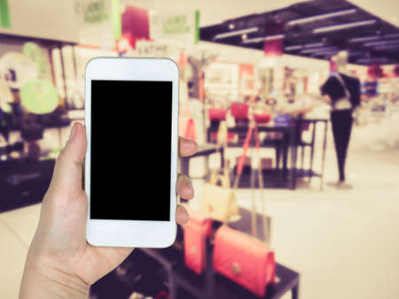Hand holding mobile smart phone on fashion shoes and bags shopping mall store on blur backgroundの写真素材