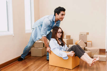 Happy couple having fun and moving woman sitting in a cardboard box at homeの写真素材
