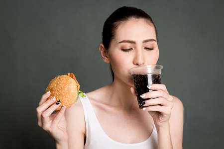 Young woman drink cola and holding hamburger on gray background. Junk food and fast food conceptの写真素材