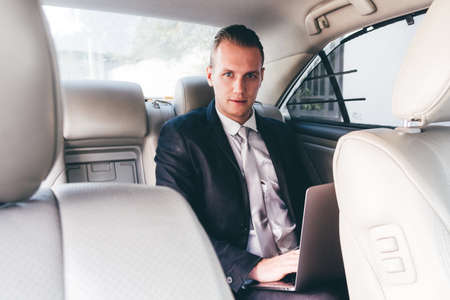 Handsome businessman working on laptop computer sitting on back seat of a carの写真素材