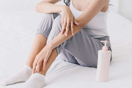 Beautiful woman sitting on bed and touching her skin for applying creamの写真素材