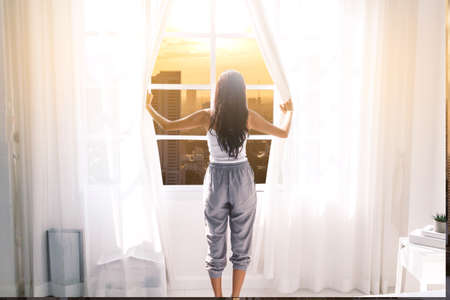 Woman waking up and opening window curtains at homeの写真素材