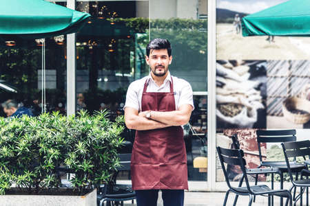 Portrait of handsome small business owner smiling and standing with crossed arms outside the cafe or coffee shop.Male barista standing at cafeのeditorial素材
