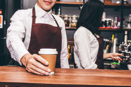 Handsome barista standing behind a counter and giving coffee cup to customer at cafeの写真素材