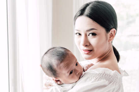 Mother holding baby in her arms in a white bedroom.Love of family conceptの写真素材