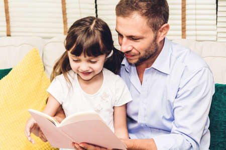Father with little daughter having fun and read the book together on the sofa at home.Little girl and daddy enjoying with story book.Love of family and conceptの写真素材