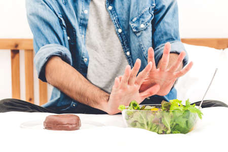 Man making choice between healthy salad and calorie bomb chocolate donut.Man rejecting the plate with healthy salad on bed at home.Healthy eating diet and Junk food conceptの写真素材