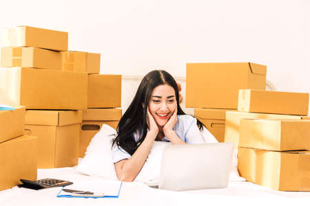 Young woman freelancer working and use laptop computer with cardboard box on bed at home - SME business online and delivery conceptの写真素材
