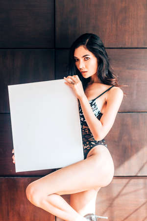 Portrait of fashion model sexy women in black bra posing and showing empty white blank board with copy space for text or ad.Advertising pretty woman naked brunetteの写真素材