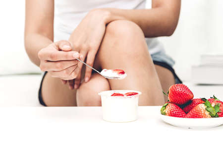 Woman eating yogurt with fresh strawberry while relaxing on sofa at home.dieting concept.healthy lifestyleの写真素材