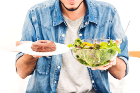 Man holding and making choice between healthy salad and calorie bomb chocolate donut on bed at home.Healthy eating and Junk food conceptの写真素材