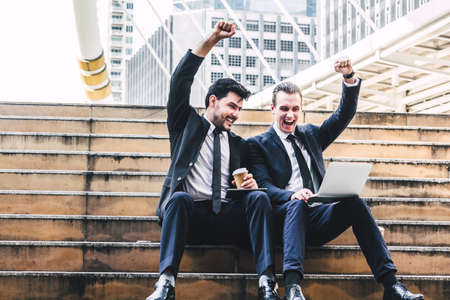 Successful of two businessman in black elegant suit sitting celebrating with arms up and working with laptop computer on the city.Business and success conceptの写真素材