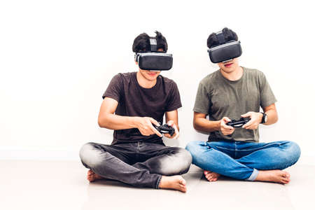 Two friends sitting and enjoying play video game with technology virtual reality together on white wall background at homeの写真素材