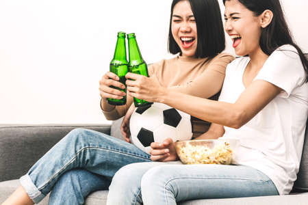 Two woman friends eating popcorn and drinking beer together and watching soccer game on sofa at home.Friendship and party conceptの写真素材