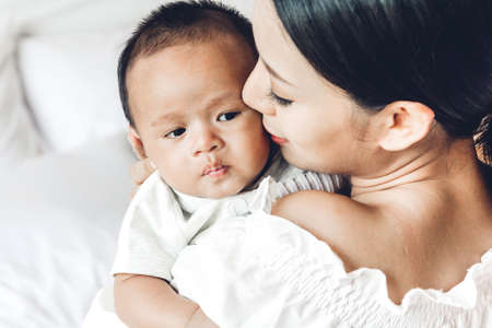 Mother kiss and holding baby in her arms  in a white bedroom.Love of family conceptの写真素材