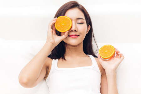 Beautiful woman holding orange slice hiding eye behind on sofaat homeの写真素材