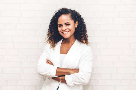 Smiling beautiful professional business african american black woman with arms crossed standing near white brick wall backgroundの写真素材