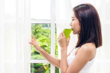 Woman enjoy healthy drinking green detox vegetable juice near window curtains in the living room at home.dieting concept.healthy lifestyleの写真素材