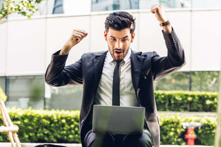 Successful of handsome businessman in black elegant suit sitting celebrating with arms up and working with laptop computer on the city.Business and startupの写真素材