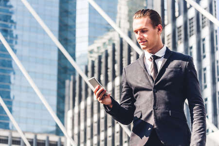 Portrait of handsome businessman in black suit standing and using smartphone outdoors on modern city background.man working with technology devices conceptの写真素材