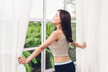 Woman waking up and opening window curtains and she feeling happy and relaxed breathing fresh air at morningの写真素材