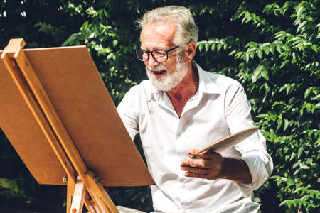 Happy smiling elder senior man artist drawing on canvas with color palette and watercolor paints in...