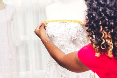 Smiling african american black woman fashion designer standing  working and using tape meter fitting on dress on the mannequin at workshop studioの写真素材
