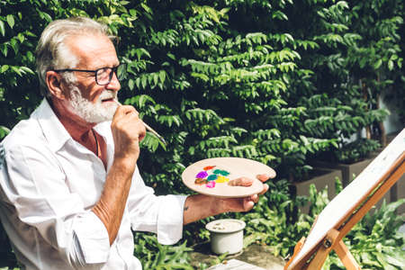 Happy smiling elder senior man artist drawing on canvas with color palette and watercolor paints in...