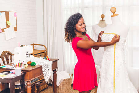 Smiling african american black woman fashion designer standing  working and using tape meter fitting on dress on the mannequin at workshop studioの写真素材