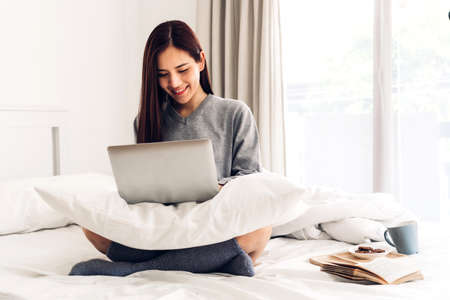 Young woman relaxing and drinking cup of hot coffee or tea using laptop computer on a cold winter day in the bedroom.woman checking social apps and working.Communication and technology conceptの写真素材
