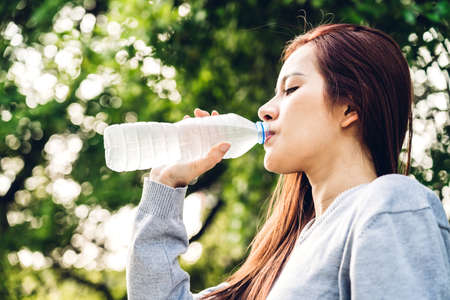 Beautiful asia woman drinking water from a bottle while relaxing and feeling fresh on green natural...