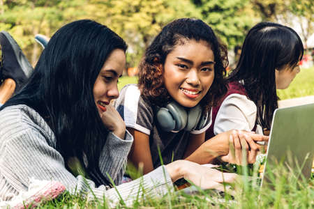 Group of smiling international students or teenagers sitting and using laptop computer doing their homework with book together in park at university.Education and friendship Conceptの写真素材
