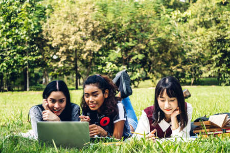 Group of smiling international students or teenagers sitting and using laptop computer doing their homework with book together in park at university.Education and friendship Concept