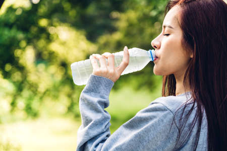 Beautiful asia woman drinking water from a bottle while relaxing and feeling fresh on green natural background at summer green park. Healthy lifestyle conceptの写真素材