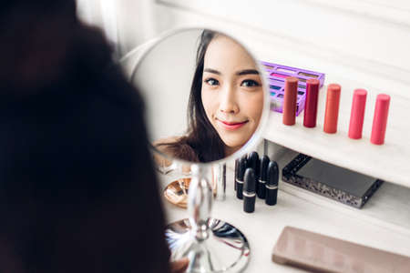 Smiling young beautiful asian woman fresh healthy skin looking on mirror with makeup cosmetics set at home.facial beauty and cosmetic conceptの写真素材