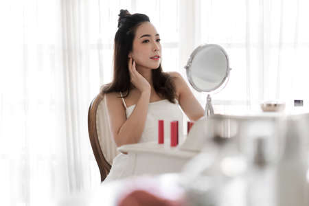 Smiling young beautiful asian woman fresh healthy skin looking on mirror with makeup cosmetics set at home.facial beauty and cosmetic conceptの写真素材