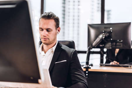Confident young handsome businessman in black elegant suit sitting and working with computer in modern officeBusiness and startup idea conceptの写真素材